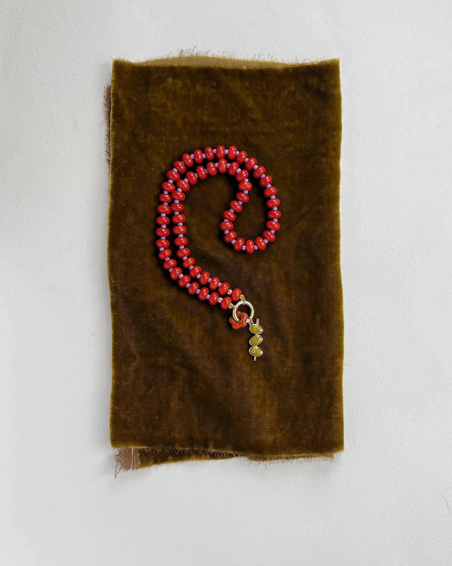 The Mod + Jo Macey Gemstone Necklace, featuring genuine red gemstones and a small gold and green charm, is elegantly displayed coiled on brown velvet atop a light gray surface.