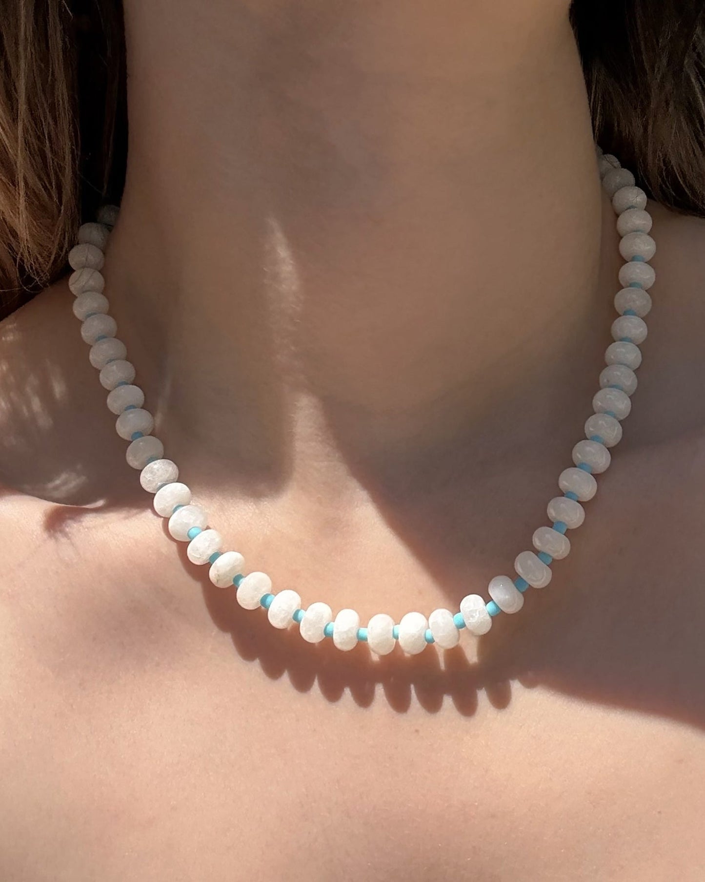 A close-up of a person’s neck wearing the Mod + Jo Macey Gemstone Necklace, featuring round, light blue and white translucent jade stones, with natural lighting highlighting the genuine gems and skin.