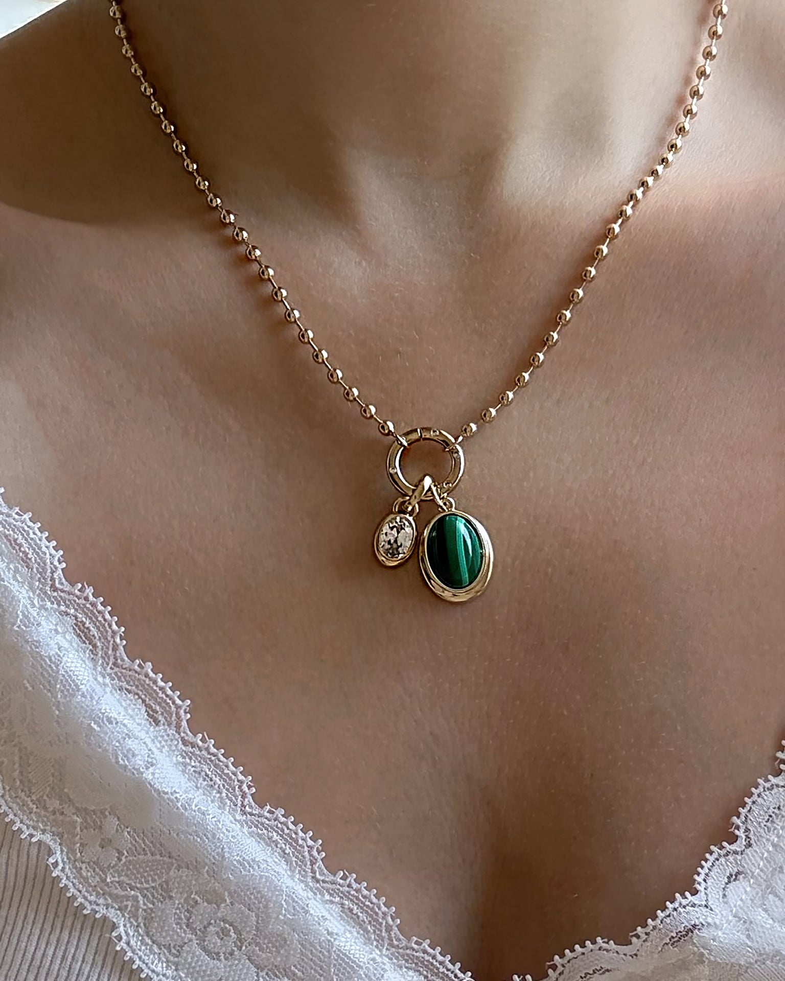 A close-up of the Mod + Jo Soren Ball Charm Clip Necklace, featuring a 14k gold ball chain with a clear gem charm and a green stone pendant, styled with a white lace-trimmed top.