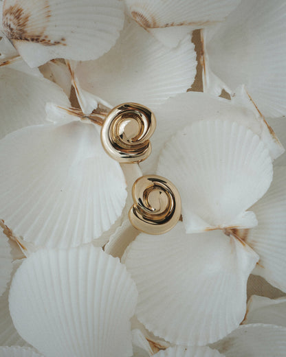 The Milley Spiral Statement Studs by Mod + Jo, crafted in shiny 14k gold, are displayed atop overlapping white scallop seashells with light brown edges.