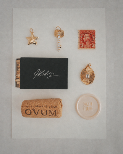 A flat lay featuring the Mod + Jo Noah Key Charm, alongside a red stamp, a black matchbox, a wine cork labeled OVUM, and a round beige item, all neatly arranged on a white background.