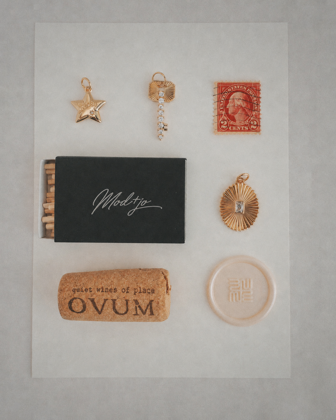 A flat lay showcases 14k gold jewelry by Mod + Jo, including a gold star necklace with the Elara Lucky Star Charm, displayed alongside a red stamp, a black matchbox, a cork with "OVUM," and a round translucent object, all neatly arranged on white paper.
