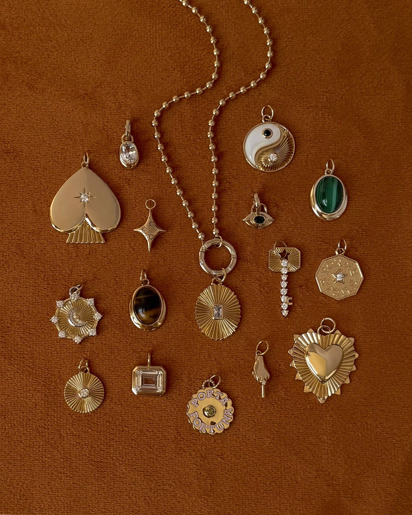 A curated selection of Mod + Jo Rory Gemstone Charm pendants—featuring heart, star, yin-yang, gemstone, and sunburst designs—are displayed on a textured brown surface. Each piece highlights the elegance of 14k gold and gold-plated craftsmanship, with some showcasing striking Tiger's Eye accents.