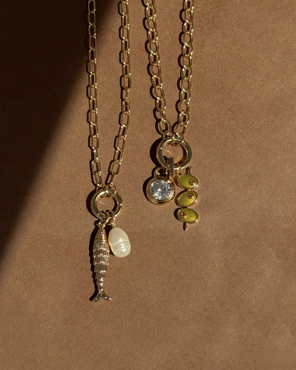 The Presley Charm Clip Necklace by Mod + Jo features two gold-plated chains: one with fish and pearl charms, the other with a round gem and green beads, both elegantly displayed on a brown surface.