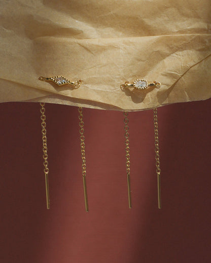 The Nicole Kite Threader Earrings by Mod + Jo, featuring delicate gold-plated chains and a sparkling cubic zirconia stone set in a gold frame at the top, hang against crumpled beige paper with a dark red background.