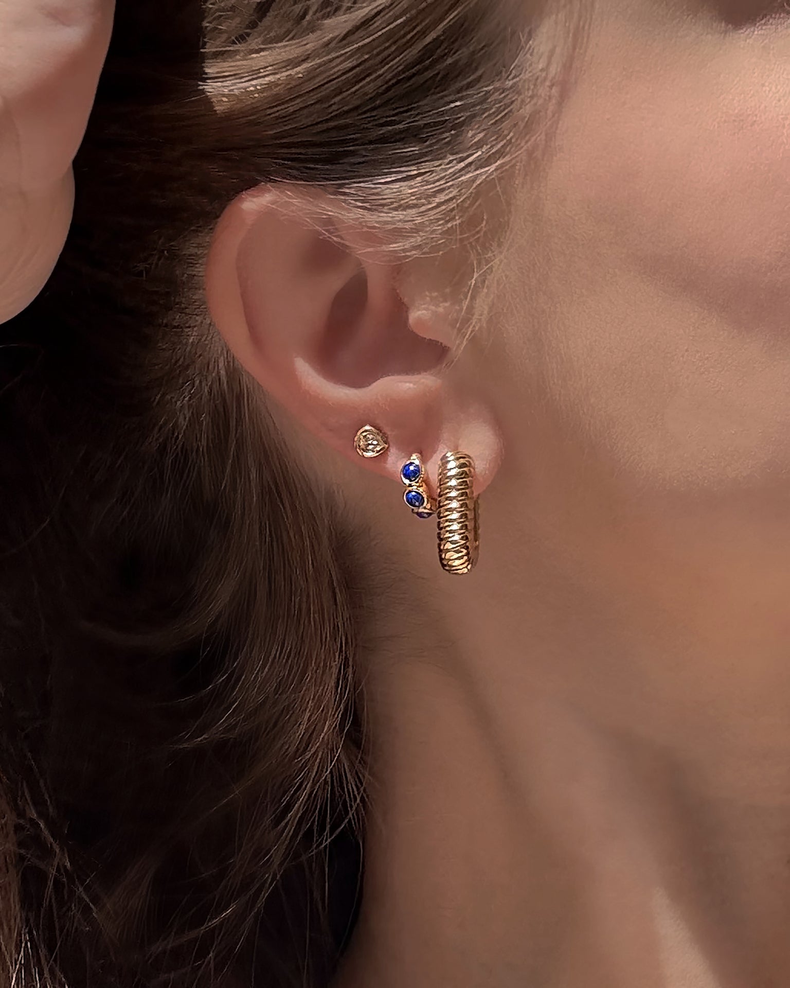 A close-up of a woman’s ear showcases three gold earrings: a simple stud, a stud featuring two blue stones, and the chunky textured Elly French Rope Hoop by Mod + Jo. Her brown hair is tucked behind her ear, drawing attention to the elegant details of her Mod + Jo gold hoop earring.