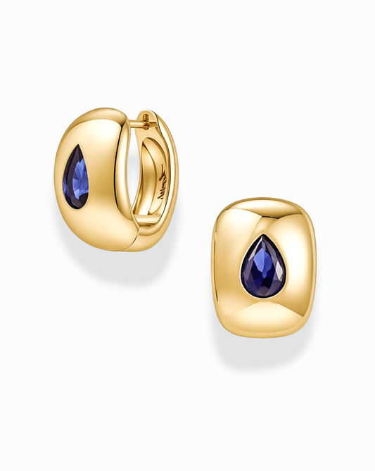 A pair of Mod + Jo Penn Gemstone Hoops in gold plated brass, designed in a rounded square huggie style and set with a teardrop-shaped blue gemstone at the center, accented by delicate CZ stones. Shown on a white background, these nickel-free earrings offer comfortable wear.