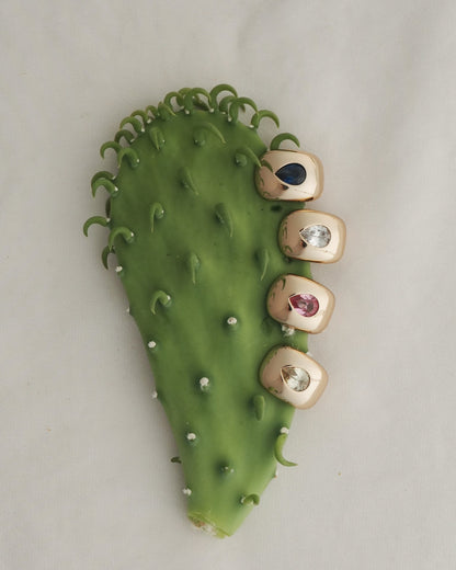 A green cactus pad displays four Mod + Jo Penn Gemstone Hoops on its right edge, each hoop adorned with different gemstones and sparkling CZ accents to resemble vibrant fingernails. The background features a soft, neutral fabric.