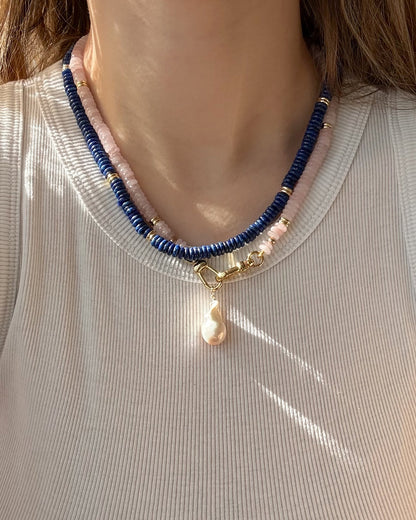 A person wearing a ribbed, light top and the Mod + Jo Sonia Gemstone Necklace—featuring two strands of lapis lazuli and pale pink beads with gold accents and a central pearl pendant.