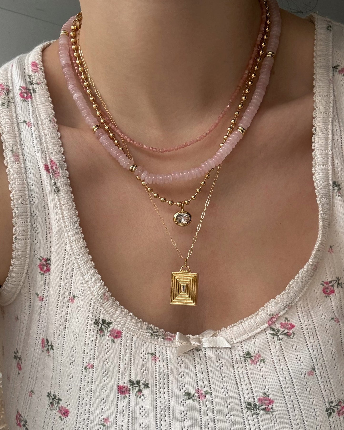 A person wears a white floral tank top with a bow, styled with pink beaded and 14k gold plated chain necklaces, plus two gold pendant necklaces—one featuring a gem, the other the Mod + Jo Inez Locket Charm.