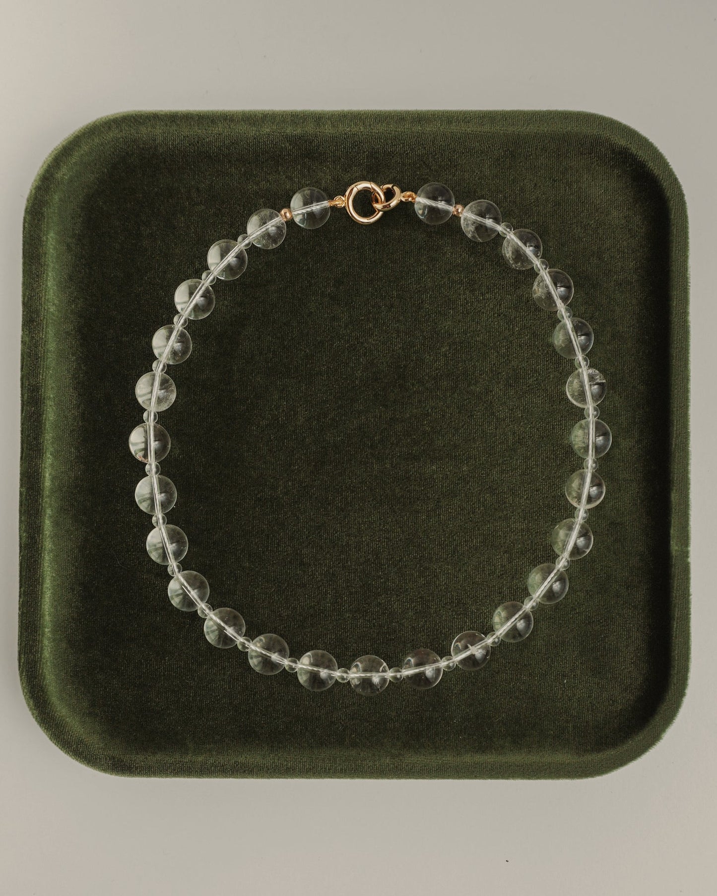 The Mod + Jo Portia Gemstone Necklace, featuring clear crystal quartz and a gold clasp, is displayed in a circle on a dark green velvet tray.