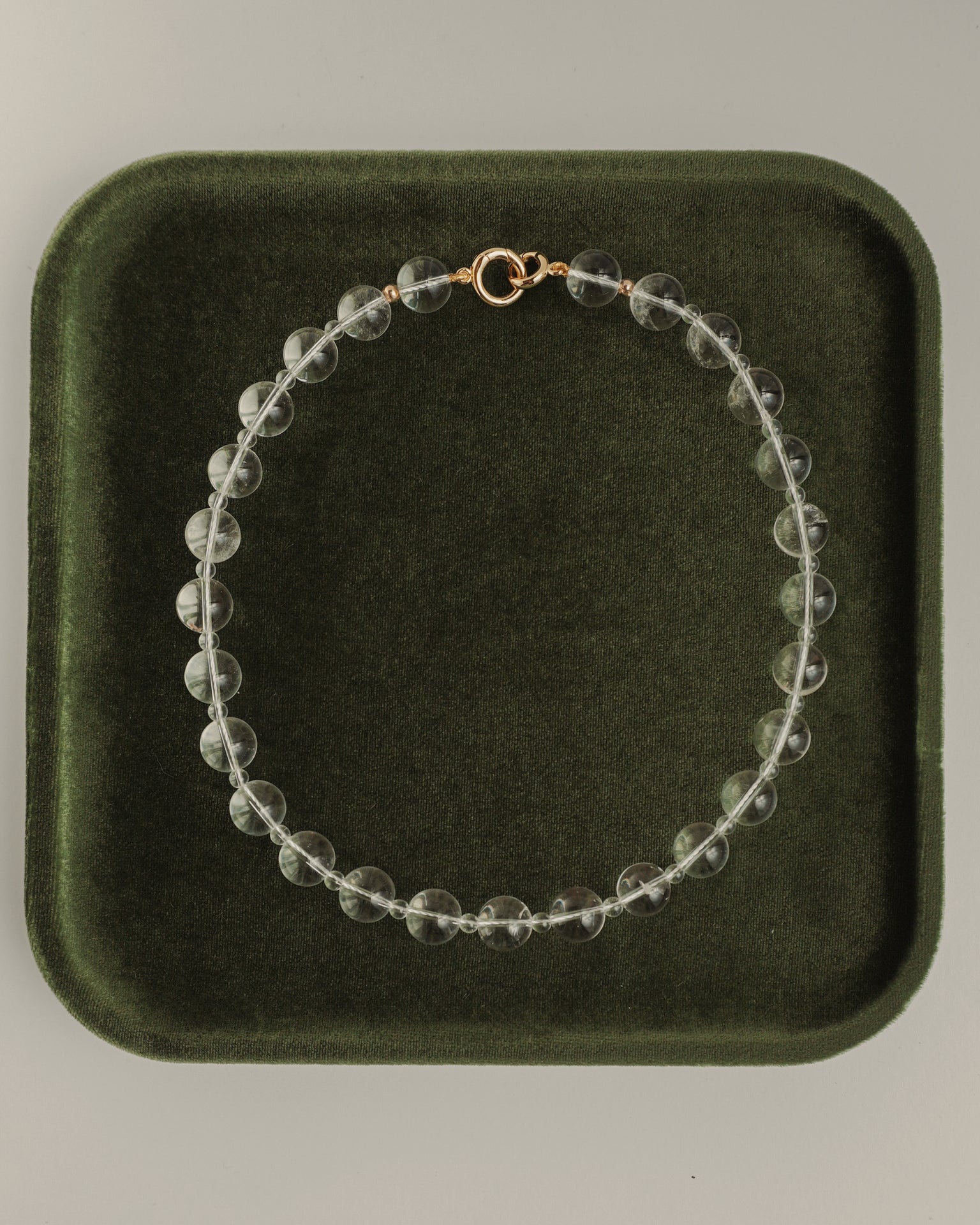 The Mod + Jo Portia Gemstone Necklace, featuring clear crystal quartz and a gold clasp, is displayed in a circle on a dark green velvet tray.
