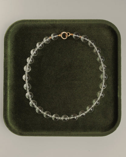 The Mod + Jo Portia Gemstone Necklace, featuring clear crystal quartz and a gold clasp, is displayed in a circle on a dark green velvet tray.