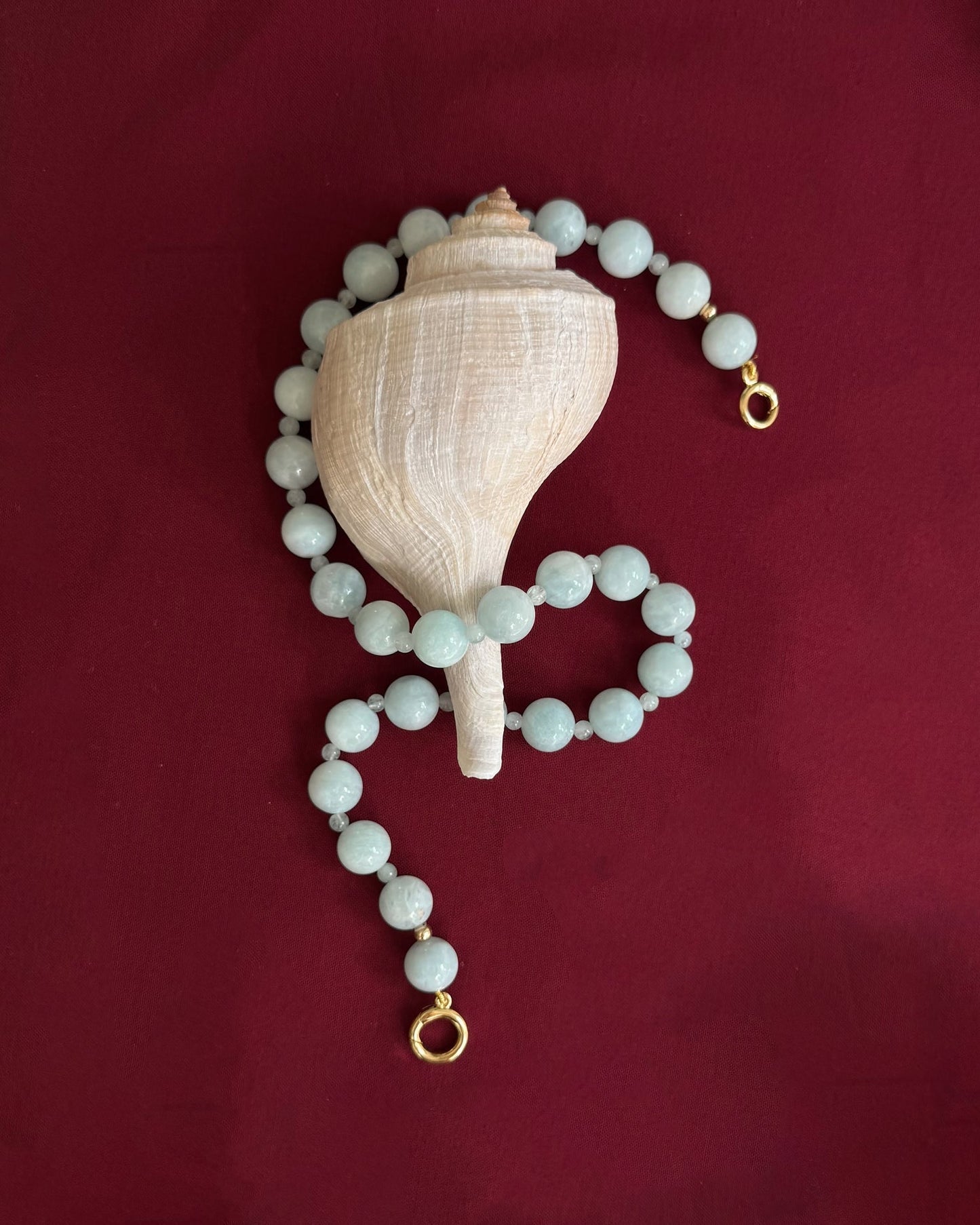 The Mod + Jo Portia Gemstone Necklace, featuring natural aquamarine beads and gold clasps, is displayed with a white conch shell on a deep red fabric backdrop.
