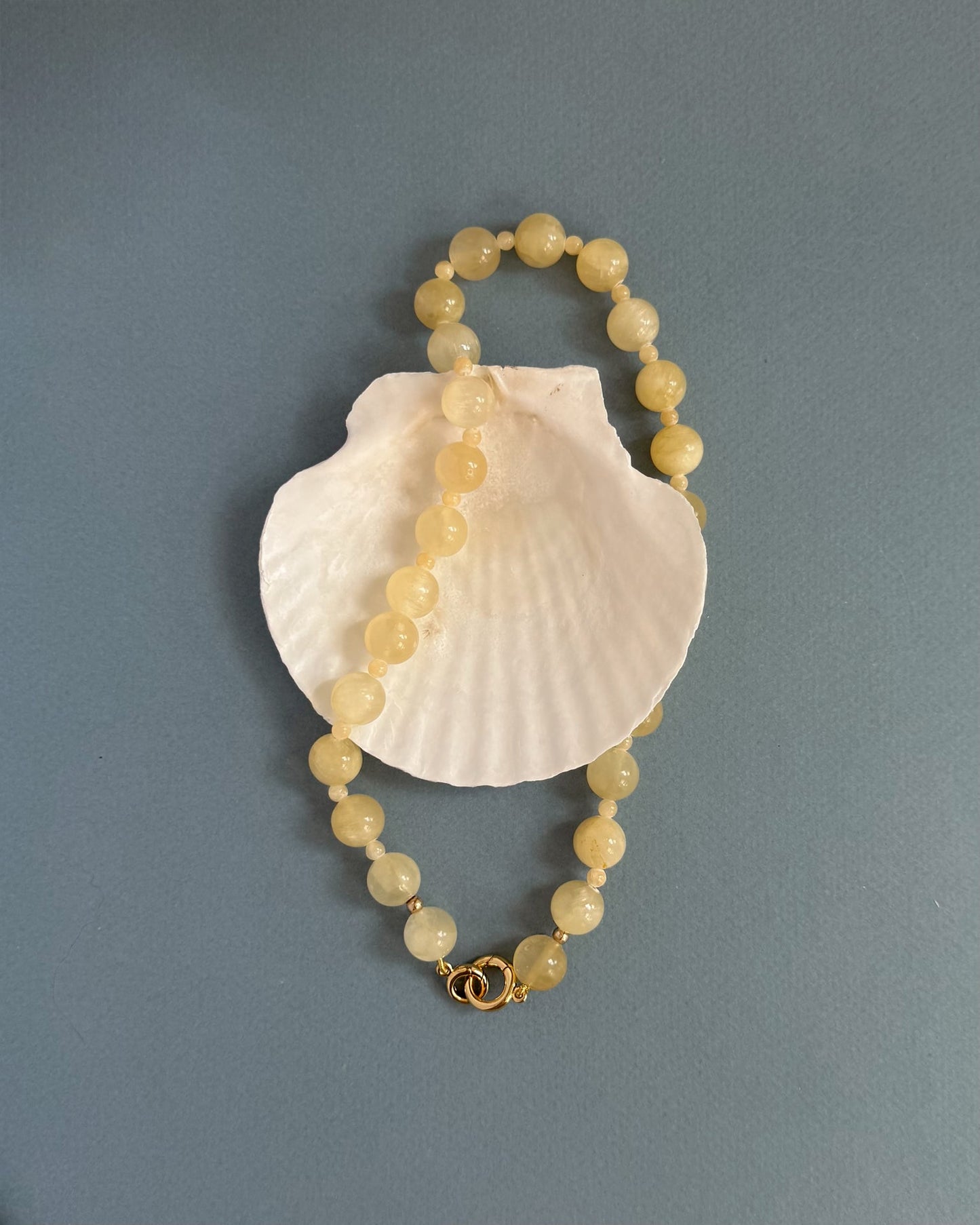 The Portia Gemstone Necklace by Mod + Jo, featuring round pale yellow beads and a gold clasp, is displayed in a loose oval atop a white seashell against a plain gray-blue background.