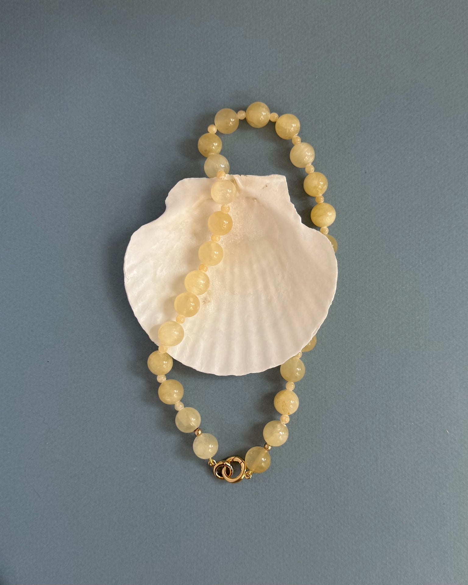The Portia Gemstone Necklace by Mod + Jo, featuring round pale yellow beads and a gold clasp, is displayed in a loose oval atop a white seashell against a plain gray-blue background.