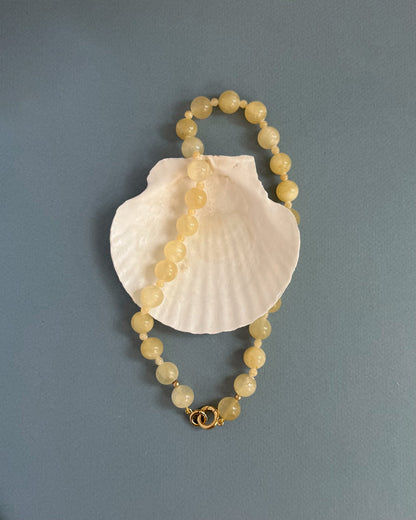 The Portia Gemstone Necklace by Mod + Jo, featuring round pale yellow beads and a gold clasp, is displayed in a loose oval atop a white seashell against a plain gray-blue background.