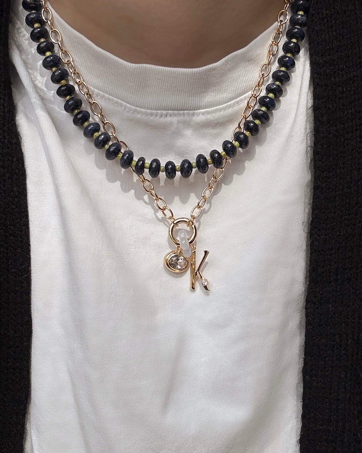A person wears the Mod + Jo Presley Charm Clip Necklace—black beads layered with a 14k gold-plated chain featuring letter and round charms—over a white shirt and black sweater.