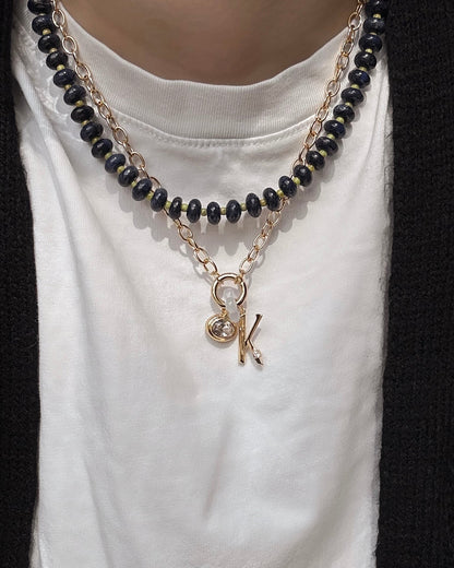 A person wears the Mod + Jo Presley Charm Clip Necklace—black beads layered with a 14k gold-plated chain featuring letter and round charms—over a white shirt and black sweater.