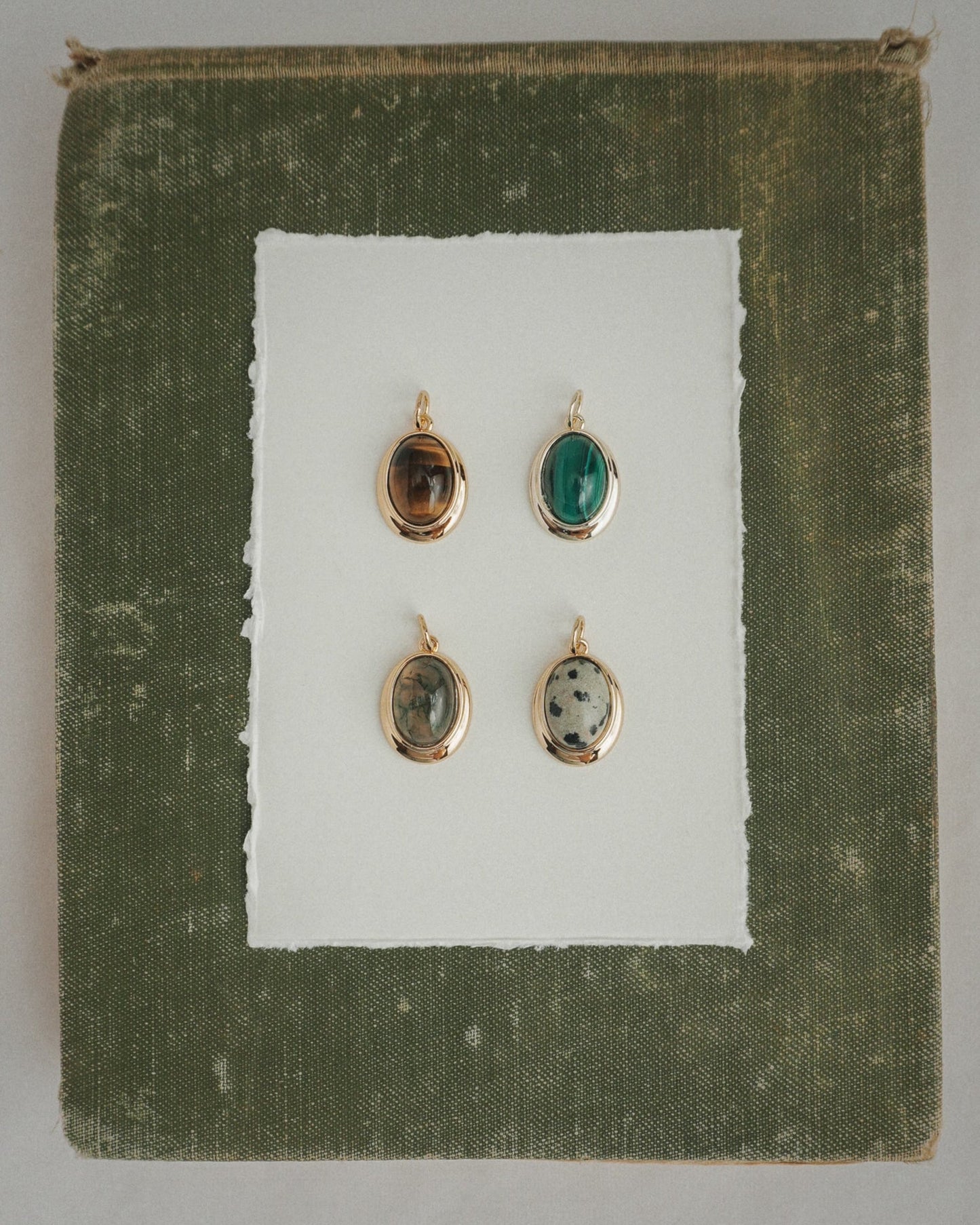 Four Rory Gemstone Charms by Mod + Jo, each featuring a unique oval gemstone with 14k gold over brass edges, are arranged on a sheet of white paper centered against a textured olive green square background. Each charm showcases a different stone—including the striking Dalmatian Jasper—and is thoughtfully crafted for an elegant touch.