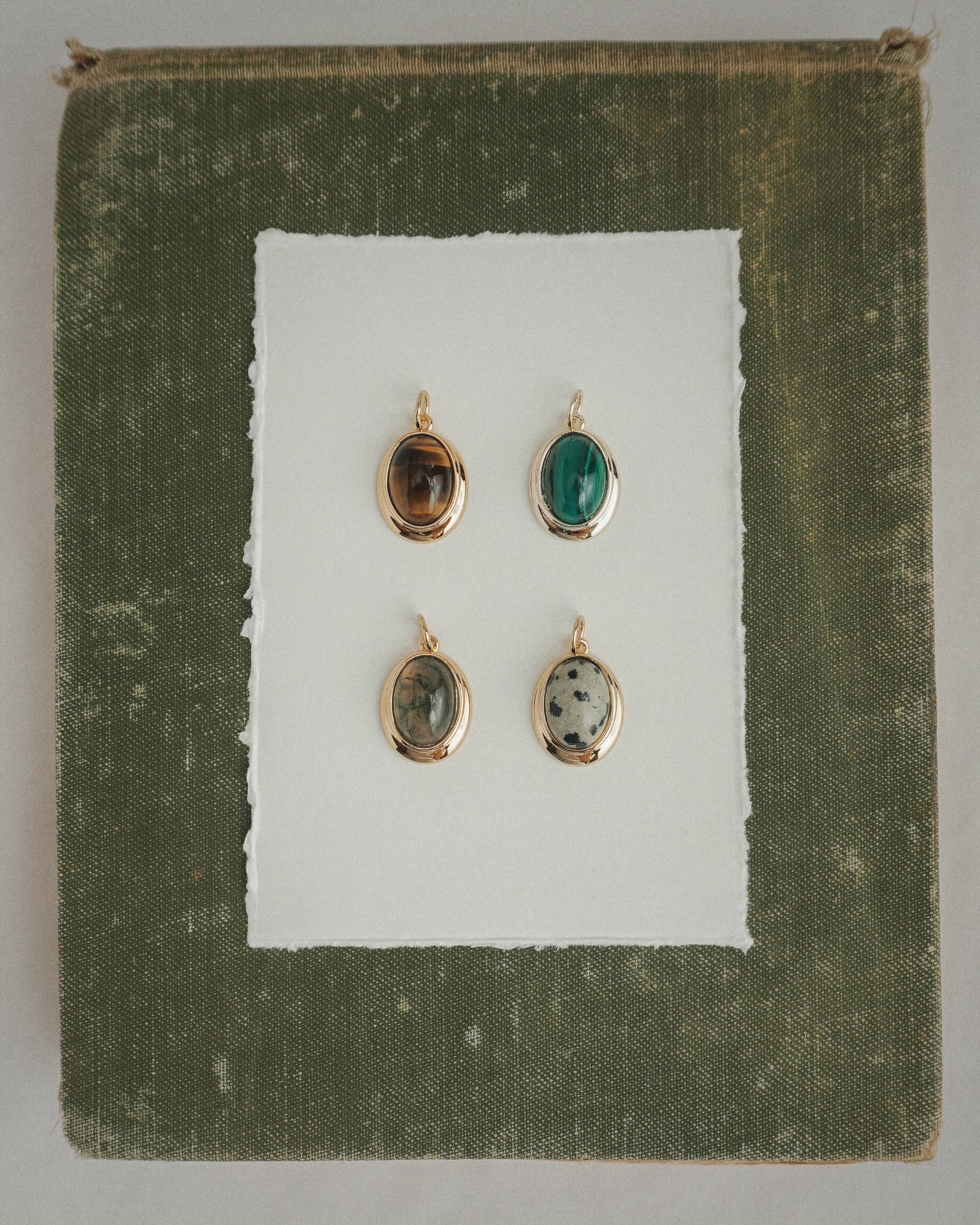 Four Rory Gemstone Charms by Mod + Jo, each featuring a unique oval gemstone with 14k gold over brass edges, are arranged on a sheet of white paper centered against a textured olive green square background. Each charm showcases a different stone—including the striking Dalmatian Jasper—and is thoughtfully crafted for an elegant touch.
