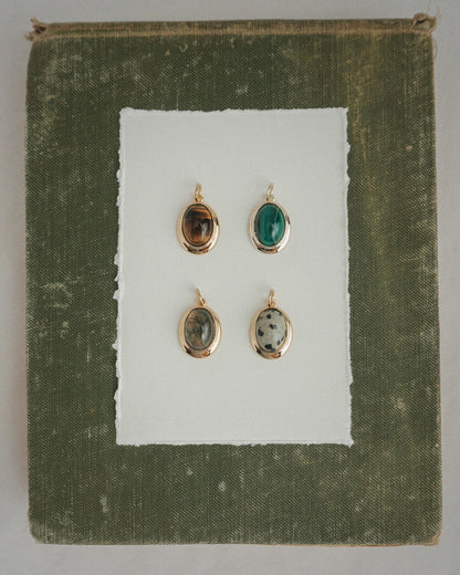Four Rory Gemstone Charms by Mod + Jo, each featuring a unique oval gemstone with 14k gold over brass edges, are arranged on a sheet of white paper centered against a textured olive green square background. Each charm showcases a different stone—including the striking Dalmatian Jasper—and is thoughtfully crafted for an elegant touch.