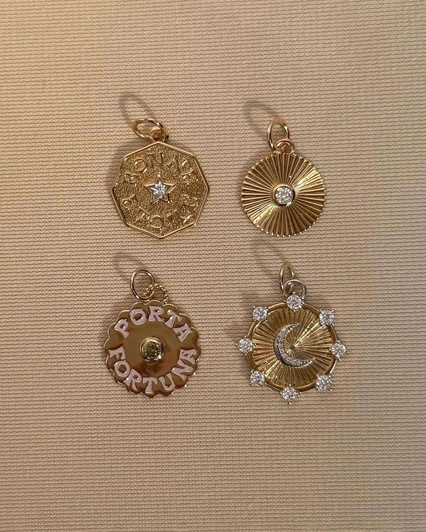 Four Mod + Jo gold pendants on a textured beige surface: the Bonne Etoile Charm, shaped as an octagon reminiscent of a lucky star necklace; a ridged circular pendant set with a gemstone; a round pendant engraved with PORTA FORTUNA in the style of gold coin jewelry; and a sunburst design adorned with a crescent moon and gemstones.
