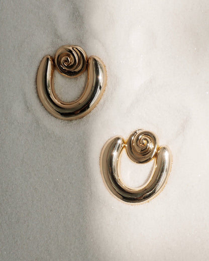The Sabine Statement Earrings by Mod + Jo feature bold 14k gold circular swirls, resting on soft white sand and catching the light—making them the perfect accessory for any occasion.