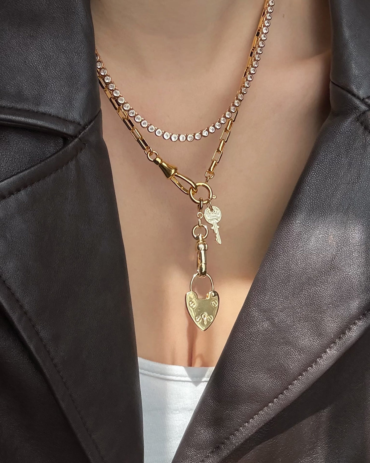 A person in a dark leather jacket and white top accessorizes with two gold necklaces: a Mod + Jo Frankie Key Charm featuring an Italian engraved key, and a chunky chain with a heart-shaped lock.