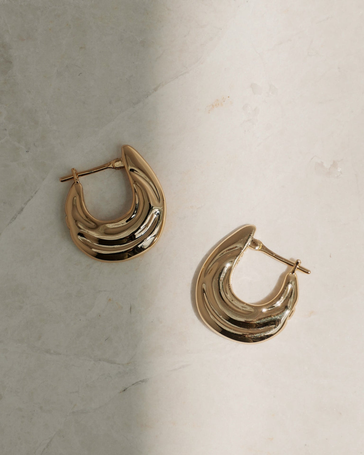 The Samara Hoops by Mod + Jo, featuring a sculpted wave design and 14k gold plating, rest on a light-colored marble surface with subtle shadows.