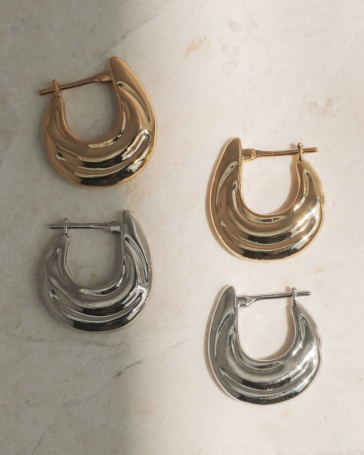 Two pairs of Samara Hoops by Mod + Jo, one gold and one silver, rest on a marble surface. Each features a crescent-shaped, layered design with advanced plating and secure latch back closures.