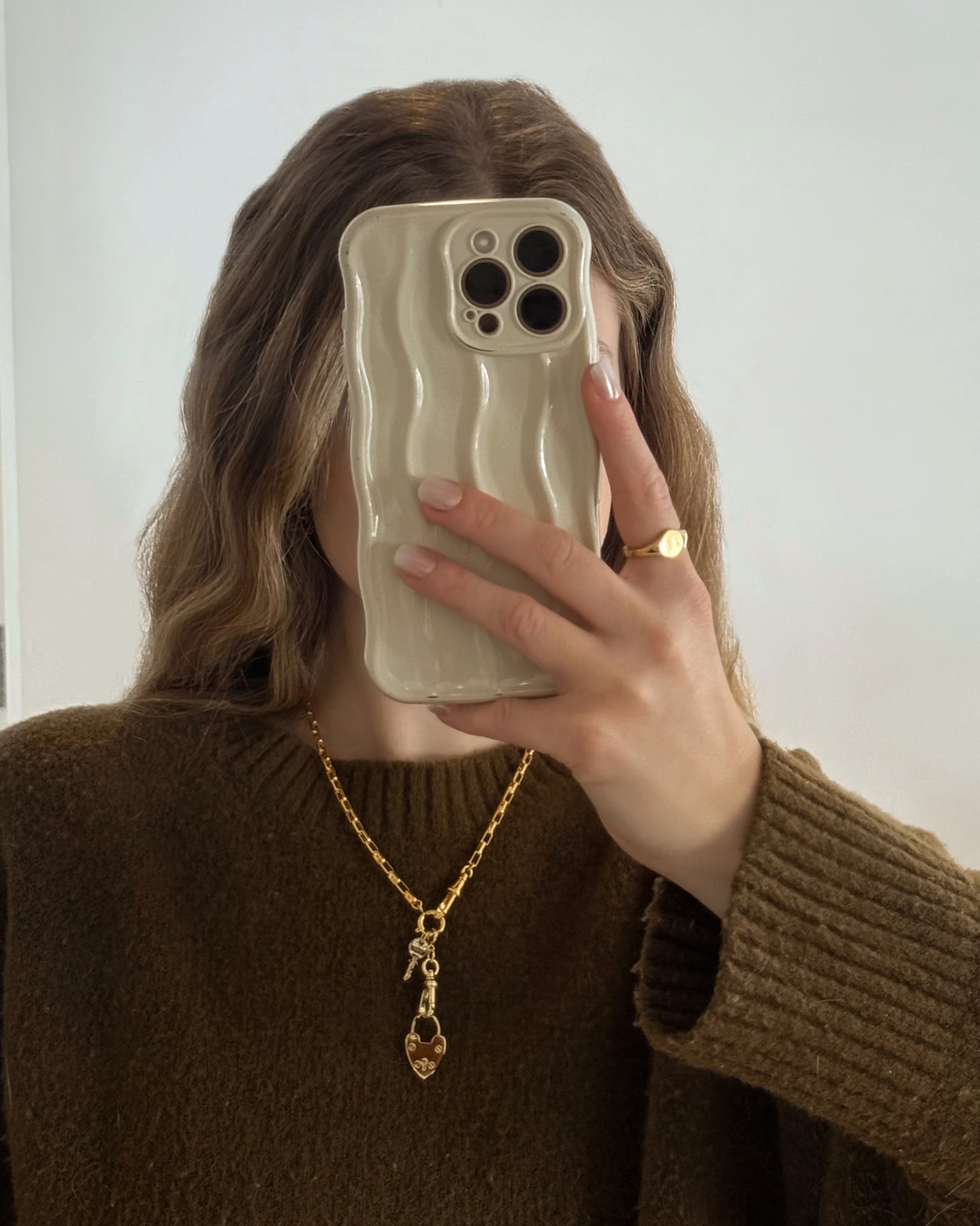 A person with wavy light brown hair wears a chunky brown sweater and Mod + Jo's Sasha Heart Locket Enhancer in 14k gold, holding a phone with a wavy beige case up to their face while taking a mirror selfie.