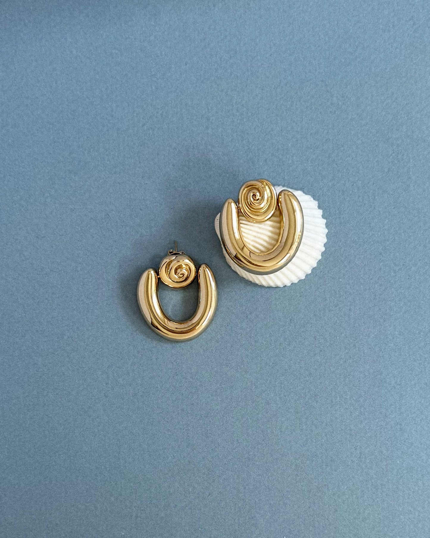 The Sabine Statement Earrings by Mod + Jo, crafted in bold 14k gold with a twisted knot design, are displayed on a grey surface—one rests atop a white seashell, while the other lies flat nearby.