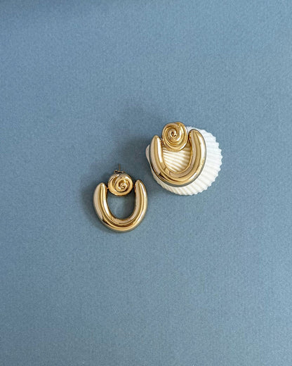 The Sabine Statement Earrings by Mod + Jo, crafted in bold 14k gold with a twisted knot design, are displayed on a grey surface—one rests atop a white seashell, while the other lies flat nearby.