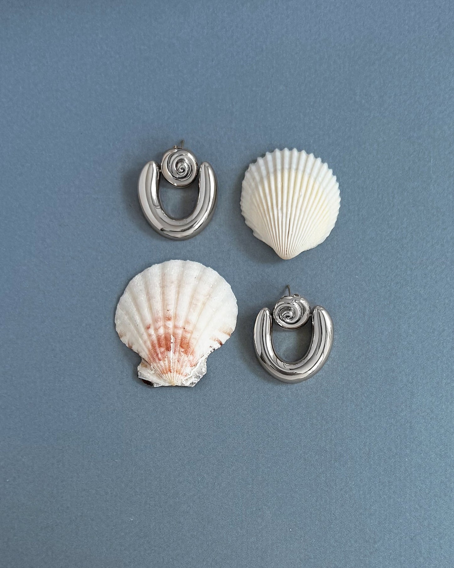 The Mod + Jo Sabine Statement Earrings in metallic gold, featuring spiral details, are displayed symmetrically on a gray-blue surface with two seashells—one white and one pinkish—for a decorative arrangement.