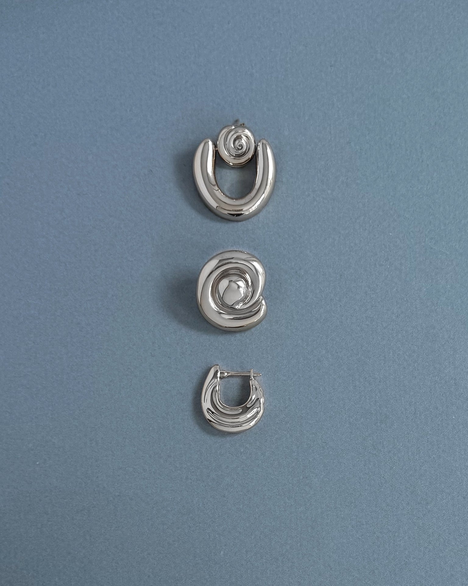 Three silver earrings from Mod + Jo are arranged vertically on a light blue textured background. The top and bottom are crescent-shaped, while the center features the Milley Spiral Statement Studs—an elegant hypoallergenic choice.