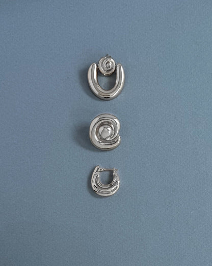 Three silver earrings from Mod + Jo are arranged vertically on a light blue textured background. The top and bottom are crescent-shaped, while the center features the Milley Spiral Statement Studs—an elegant hypoallergenic choice.