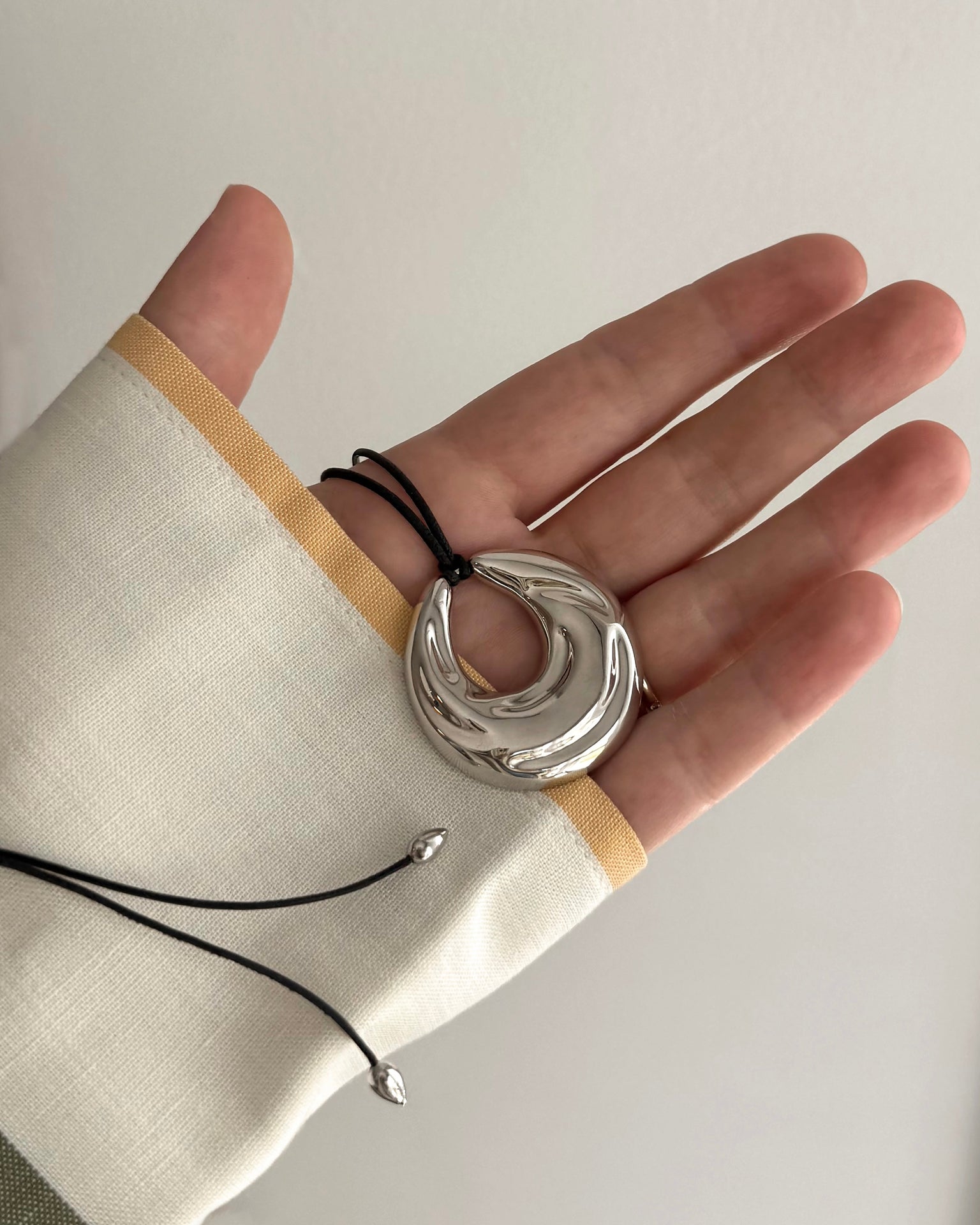 A hand displays the Mod + Jo Kira Cord Necklace, featuring a shiny abstract silver adjustable pendant on a black cord. The wrist wears a cream sleeve with tan trim and black strings, set against a neutral background.