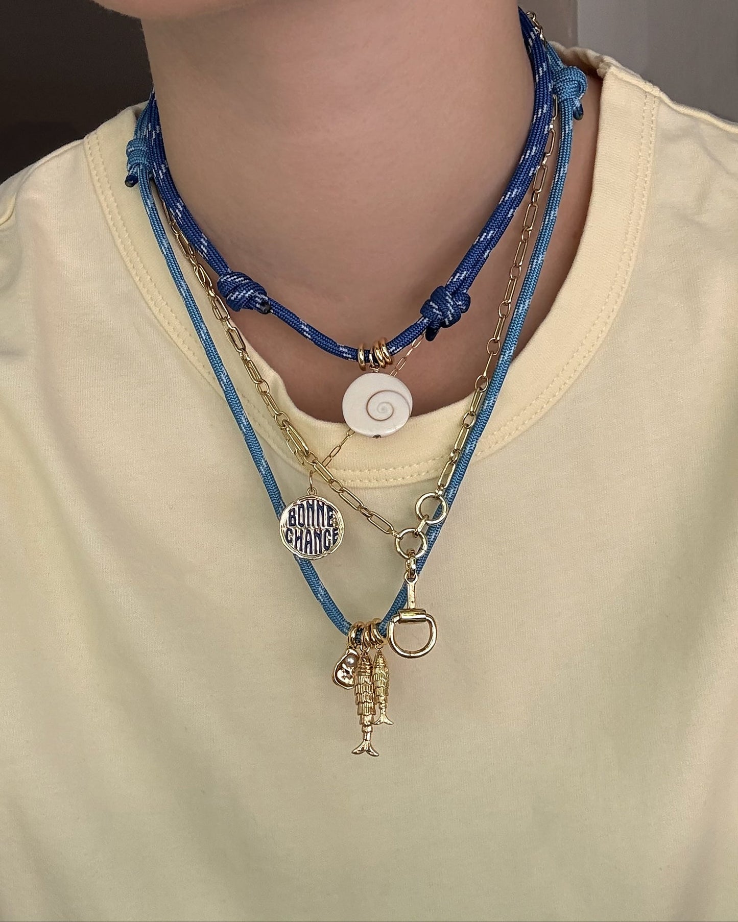 A person in a pale yellow shirt layers necklaces: the Mod + Jo Tommy Cord Necklace in blue, featuring a white spiral shell, along with two gold chains—one with a “BONNE CHANCE” charm and another with fishing lure-like pendants.