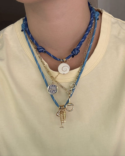 A person in a pale yellow shirt layers necklaces: the Mod + Jo Tommy Cord Necklace in blue, featuring a white spiral shell, along with two gold chains—one with a “BONNE CHANCE” charm and another with fishing lure-like pendants.