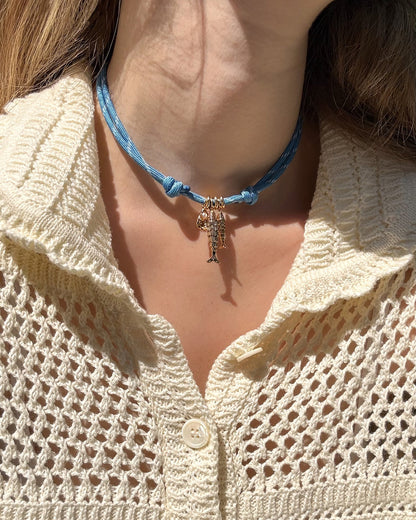 A person wearing a cream, textured knit top and the Mod + Jo Tommy Cord Necklace in blue with gold-plated spacers, photographed in sunlight.