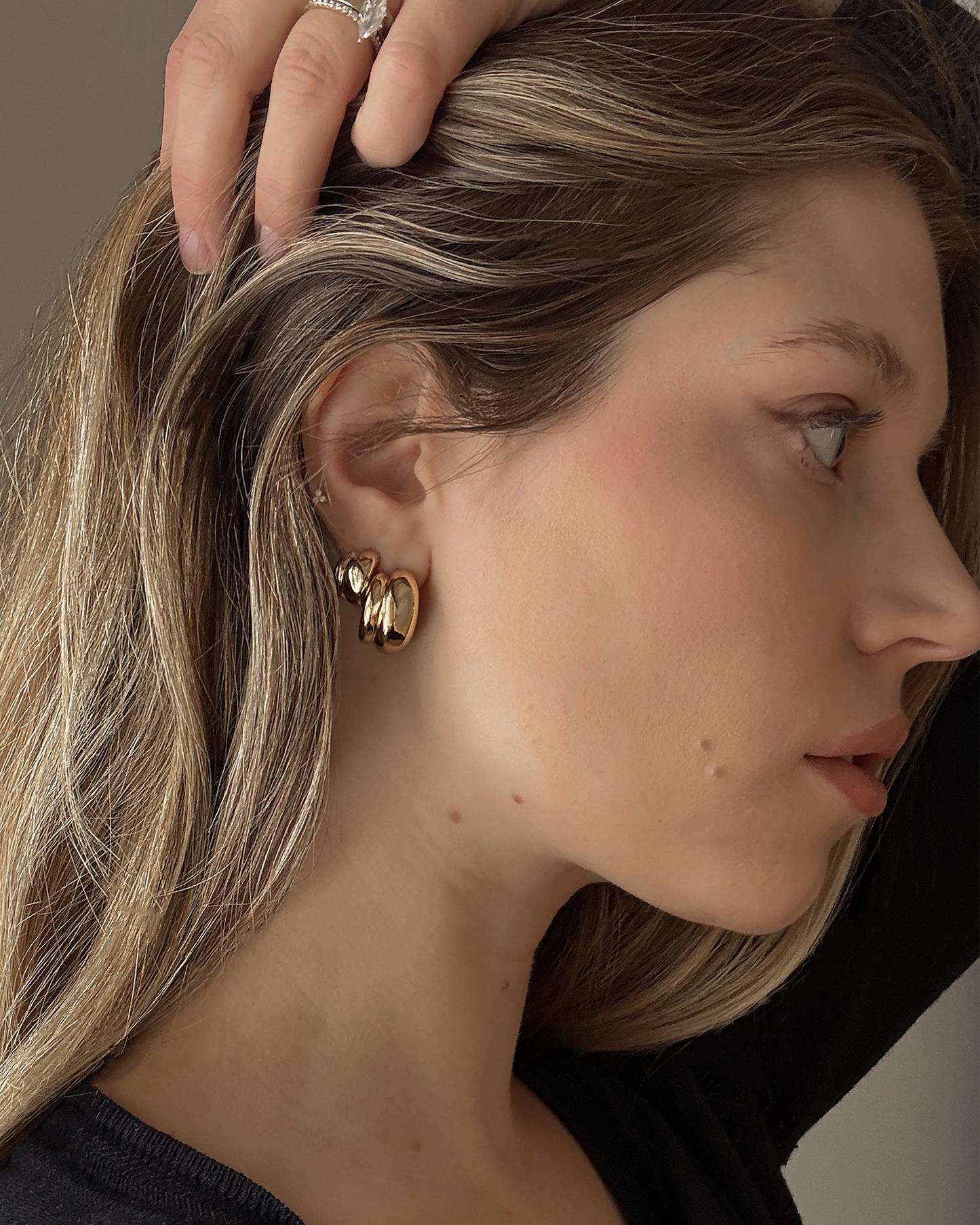 A woman with long, light brown hair wears Mod + Jo’s Grande Sofie Volume Hoops and a black top, resting her hand on her head to reveal a ring as she gazes right in soft light.