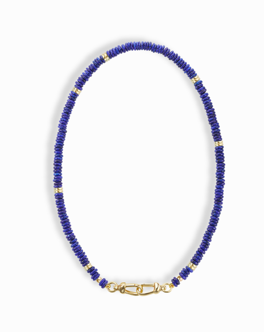 The Sonia Gemstone Necklace by Mod + Jo features small, round blue lapis lazuli beads with gold spacer beads and a gold clasp, displayed in an oval shape on a white background.