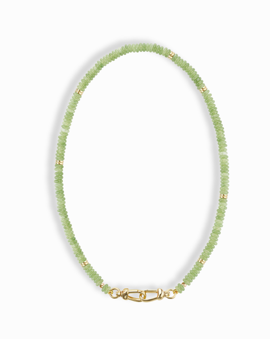 The Sonia Gemstone Necklace by Mod + Jo features small, round, light green genuine gemstone beads with gold spacers and a gold clasp, elegantly arranged in an oval shape against a white background.
