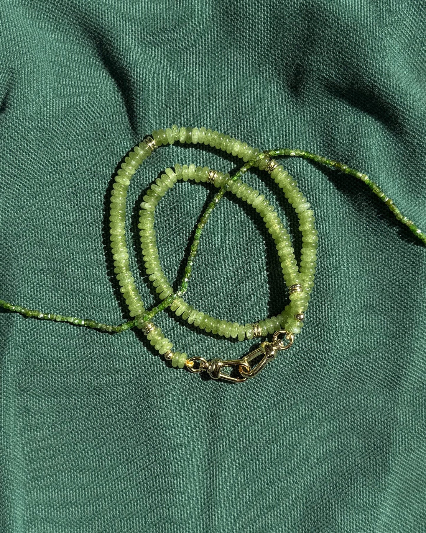 The Merit Gemstone Necklace by Mod + Jo, featuring green beads, gold accents, and a 14k gold-filled clasp, is elegantly displayed coiled on textured green fabric.