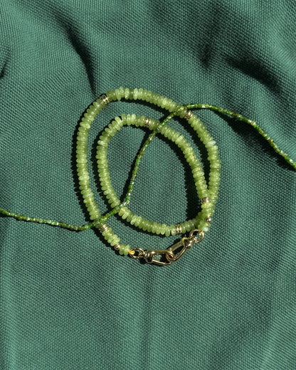 The Merit Gemstone Necklace by Mod + Jo, featuring green beads, gold accents, and a 14k gold-filled clasp, is elegantly displayed coiled on textured green fabric.