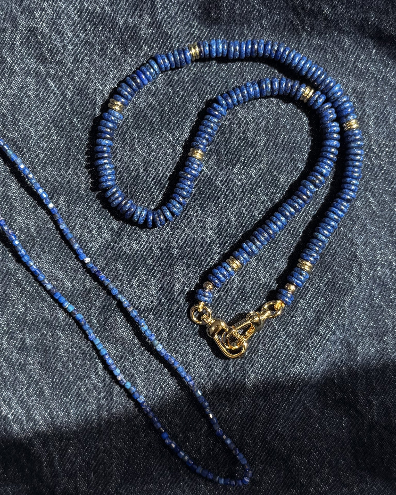 Two Mod + Jo Sonia Gemstone Necklaces, featuring lapis lazuli beads, rest on dark denim; one is thicker with gold accents and clasp, while the other is thinner with smaller, uniform beads.