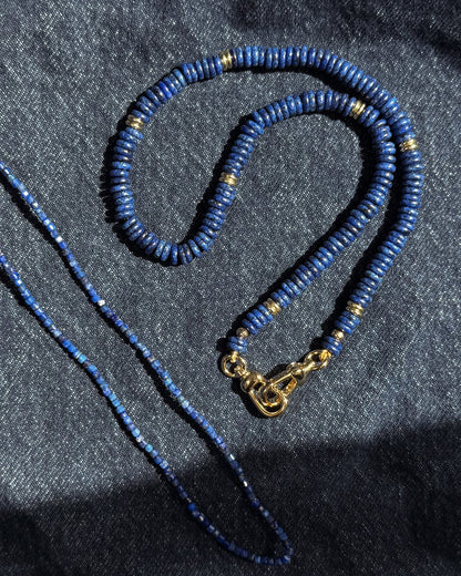 Two Mod + Jo Sonia Gemstone Necklaces, featuring lapis lazuli beads, rest on dark denim; one is thicker with gold accents and clasp, while the other is thinner with smaller, uniform beads.