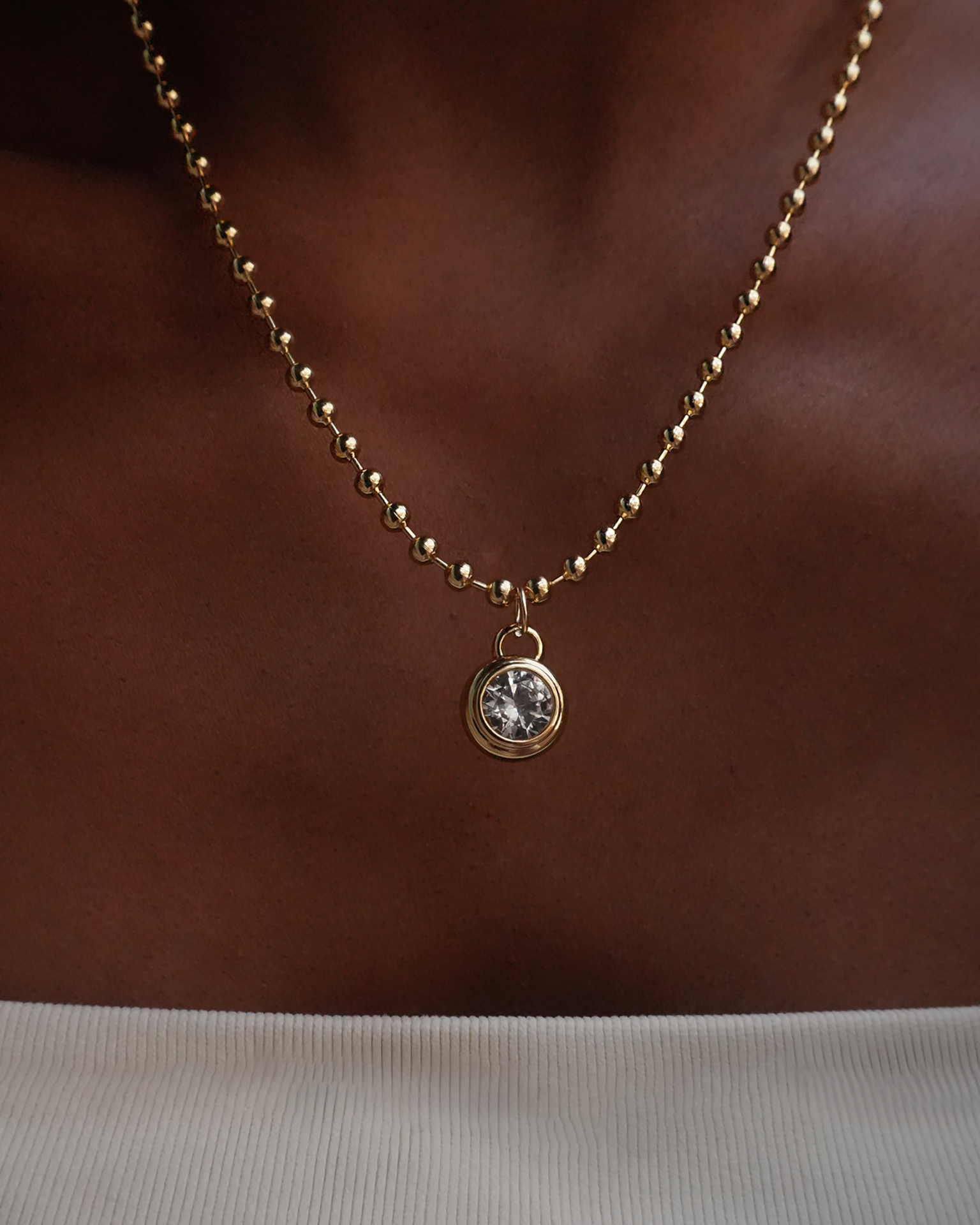 A person wearing a white ribbed top has the Mod + Jo *Preorder* Tove Sapphire Bezel Charm, a 14k gold beaded necklace with a round lab-grown sapphire pendant, around their neck.