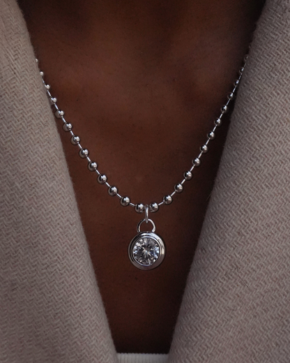 Silver ball chain necklace with a round sapphire bezel pendant on a person wearing a beige cardigan.